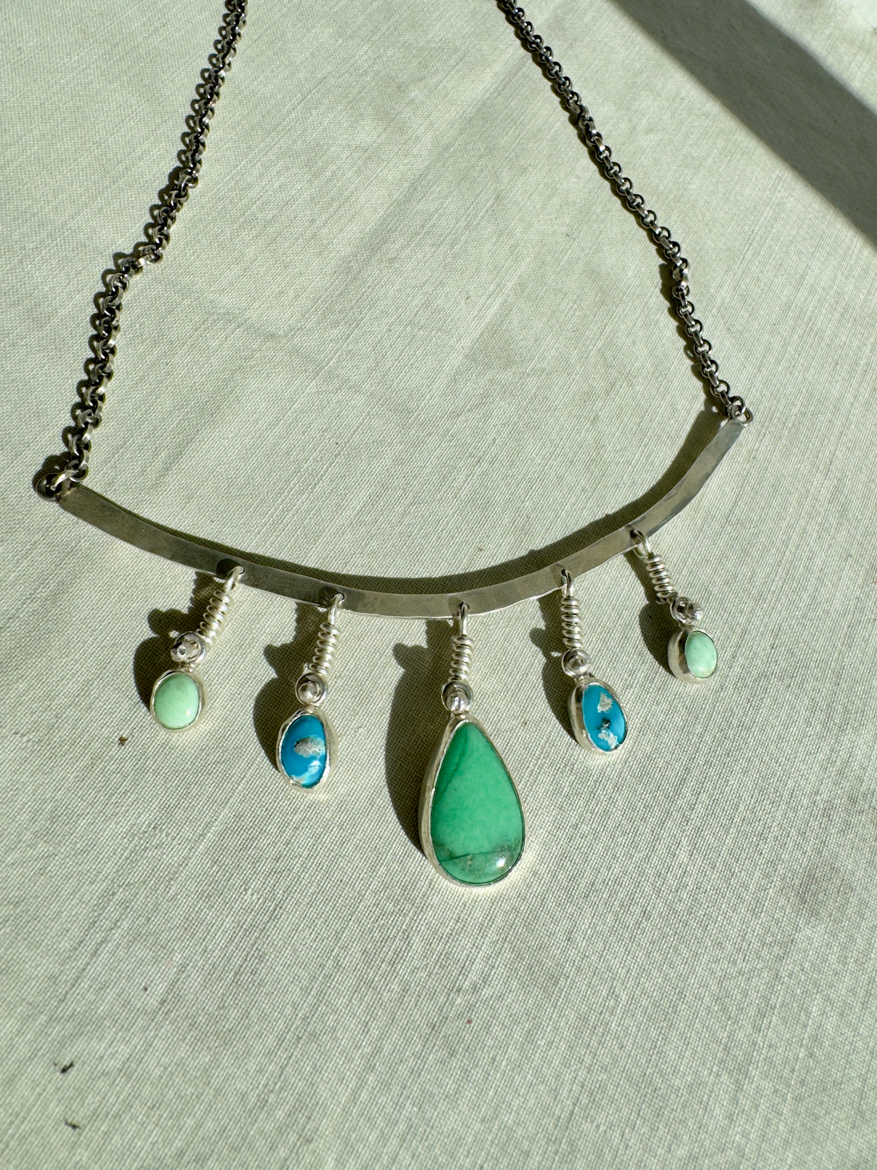 Sterling Silver hammered bar necklace with beautiful green Lucid center stone, flanked by two bright blue castle dome stones and two pale green lucin variscite stones for a total of five stones. The sterling silver bar is attached to a 2.7mm flat rolo chain, oxidized to give it a darker color, with a sterling silver toggle clasp. The necklace rests right on your collarbone, making it the perfect necklace to dress up or down.