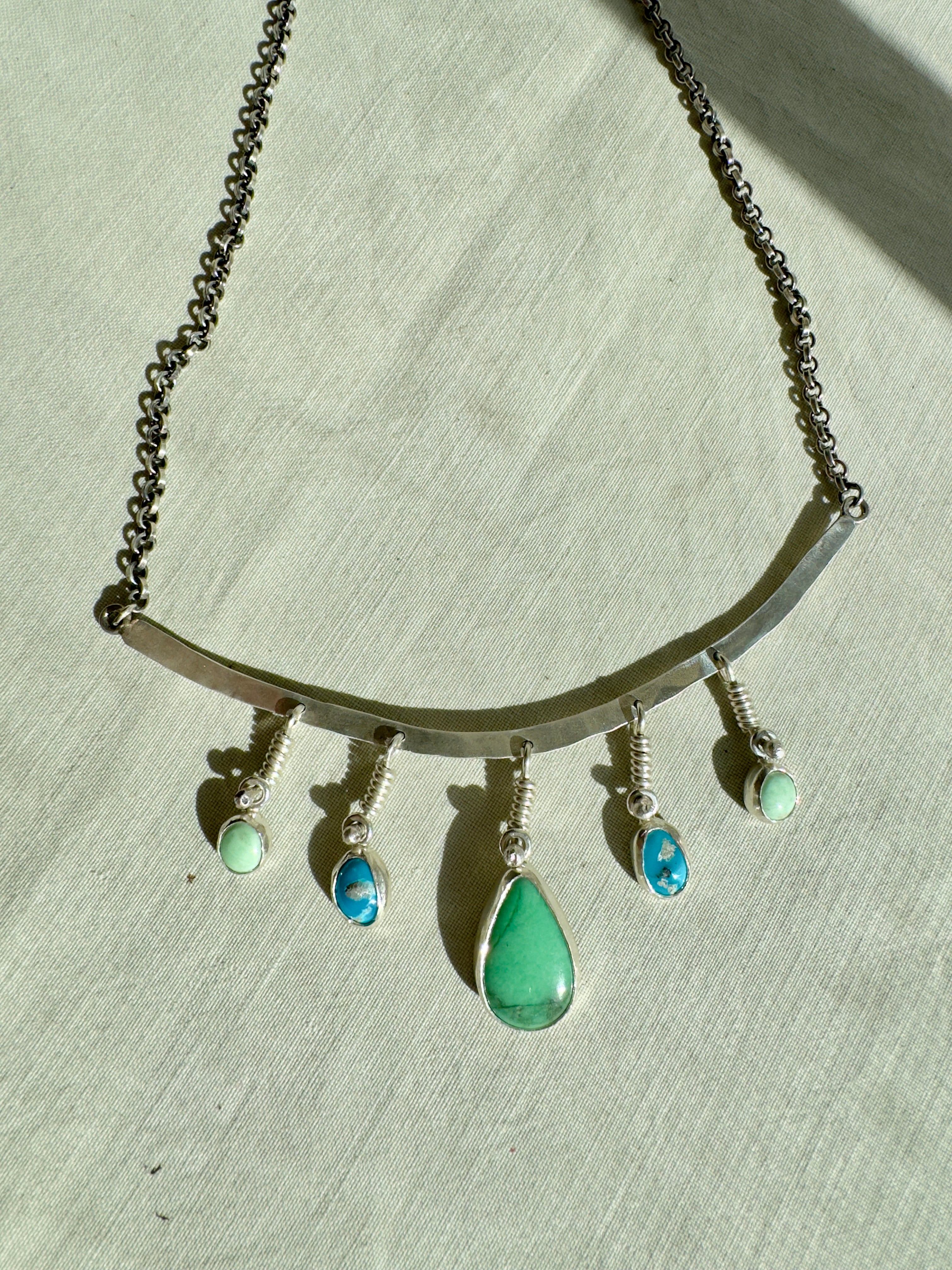 Sterling Silver hammered bar necklace with beautiful green Lucid center stone, flanked by two bright blue castle dome stones and two pale green lucin variscite stones for a total of five stones. The sterling silver bar is attached to a 2.7mm flat rolo chain, oxidized to give it a darker color, with a sterling silver toggle clasp. The necklace rests right on your collarbone, making it the perfect necklace to dress up or down.