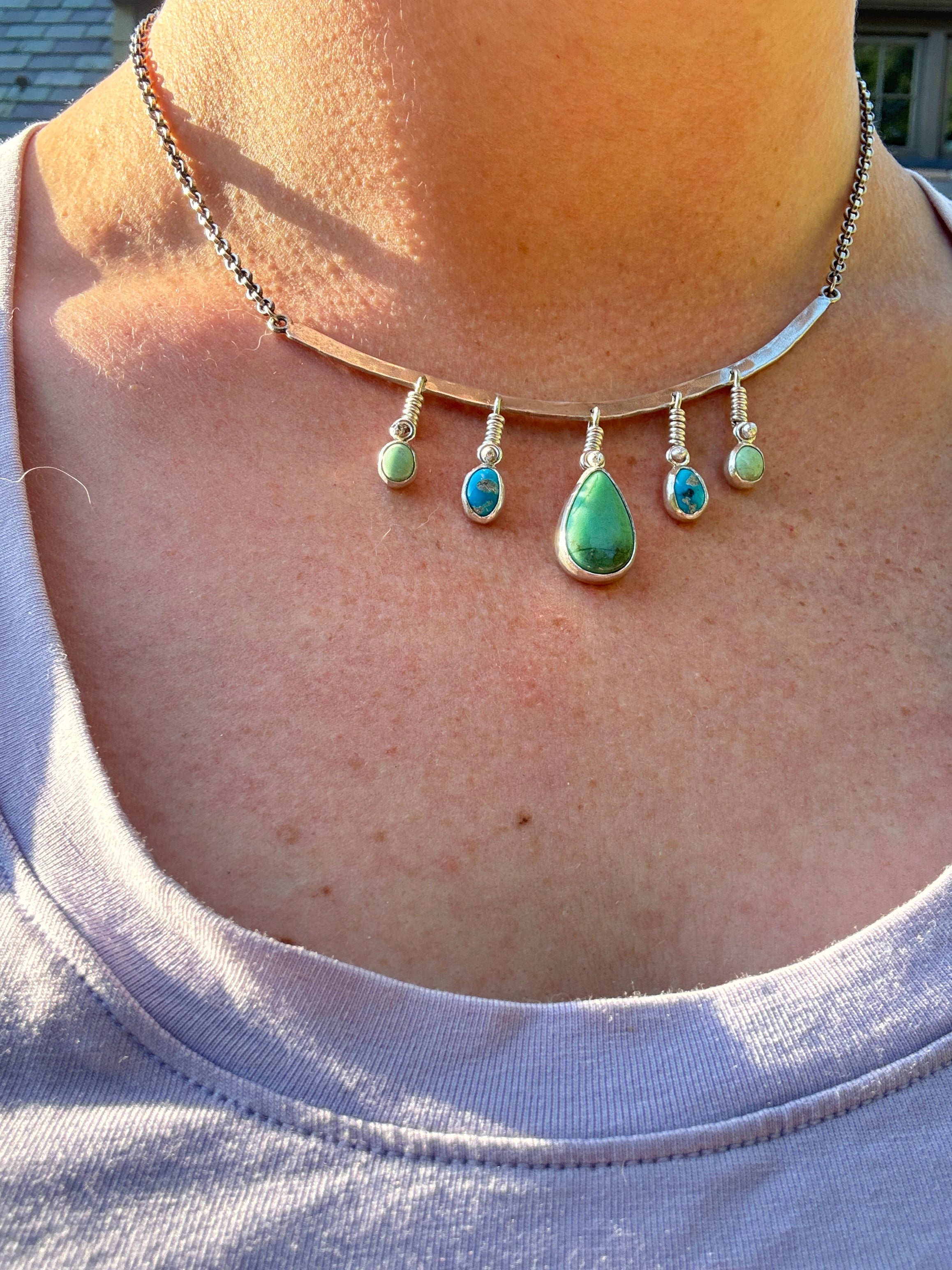 Sterling Silver hammered bar necklace with beautiful green Lucid center stone, flanked by two bright blue castle dome stones and two pale green lucin variscite stones for a total of five stones. The sterling silver bar is attached to a 2.7mm flat rolo chain, oxidized to give it a darker color, with a sterling silver toggle clasp. The necklace rests right on your collarbone, making it the perfect necklace to dress up or down.